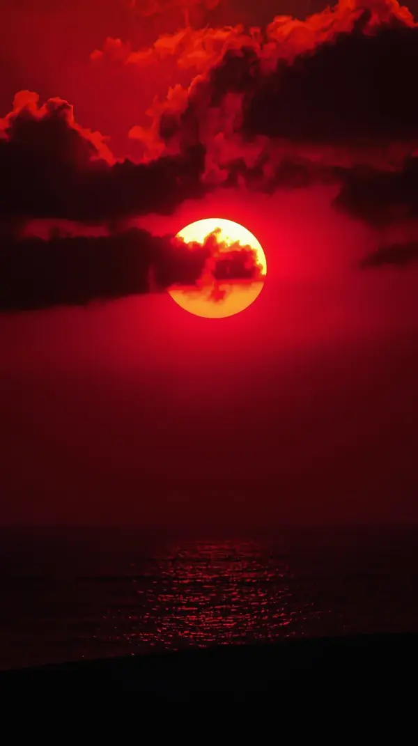 a glowing yellow sun partially hidden by dark clouds in a vibrant crimson red sky over the ocean